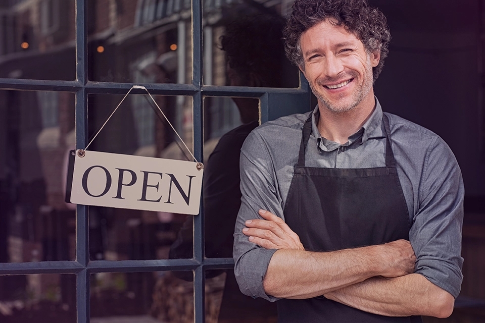 A small business owner smiling outside of the entrance of his shop, the door has an open sign. 